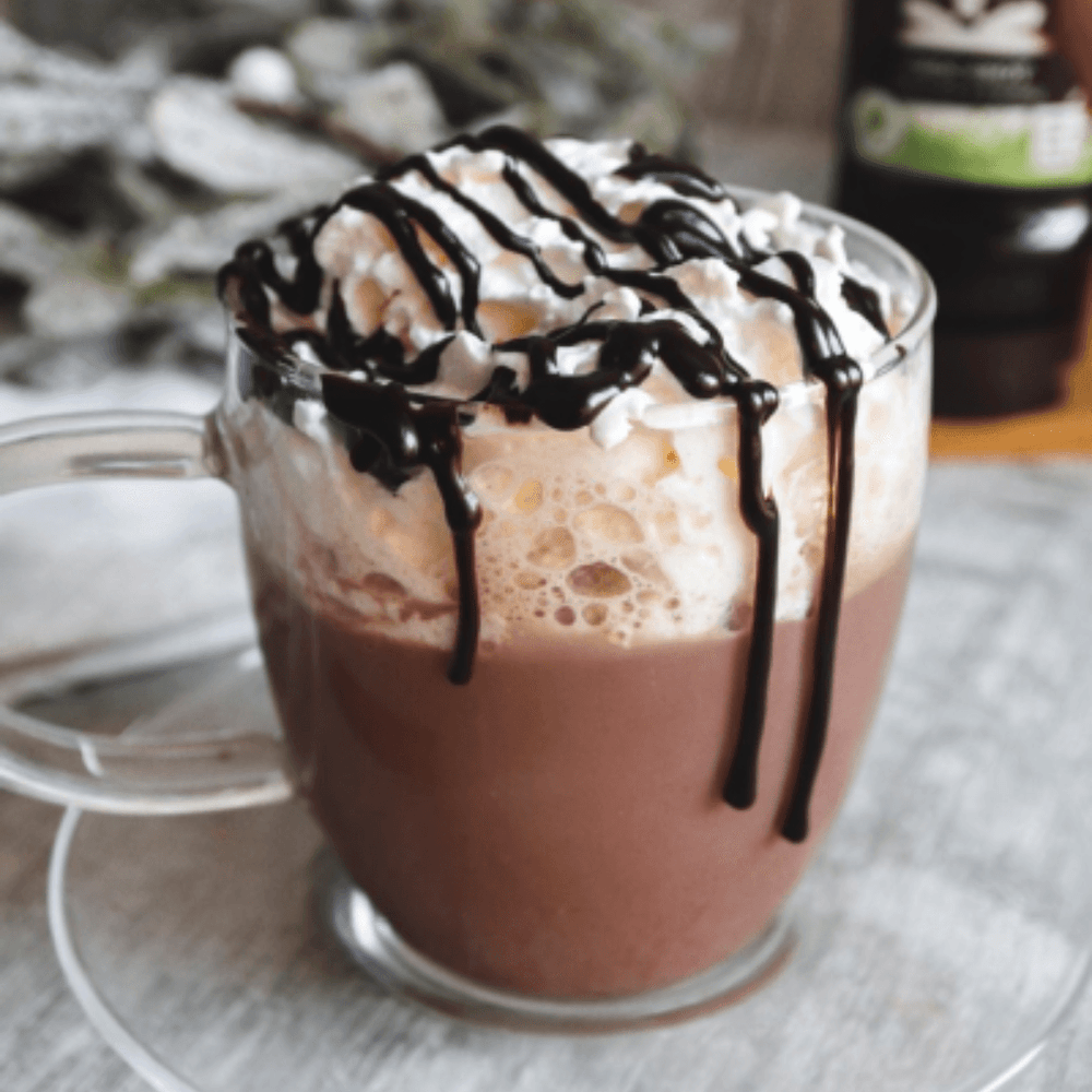 Delicious looking hot chocolate made using Sweet Freedom Choc Shot in a glass mug topped with vegan squirty cream and drizzled with more Choc Shot chocolate syrup