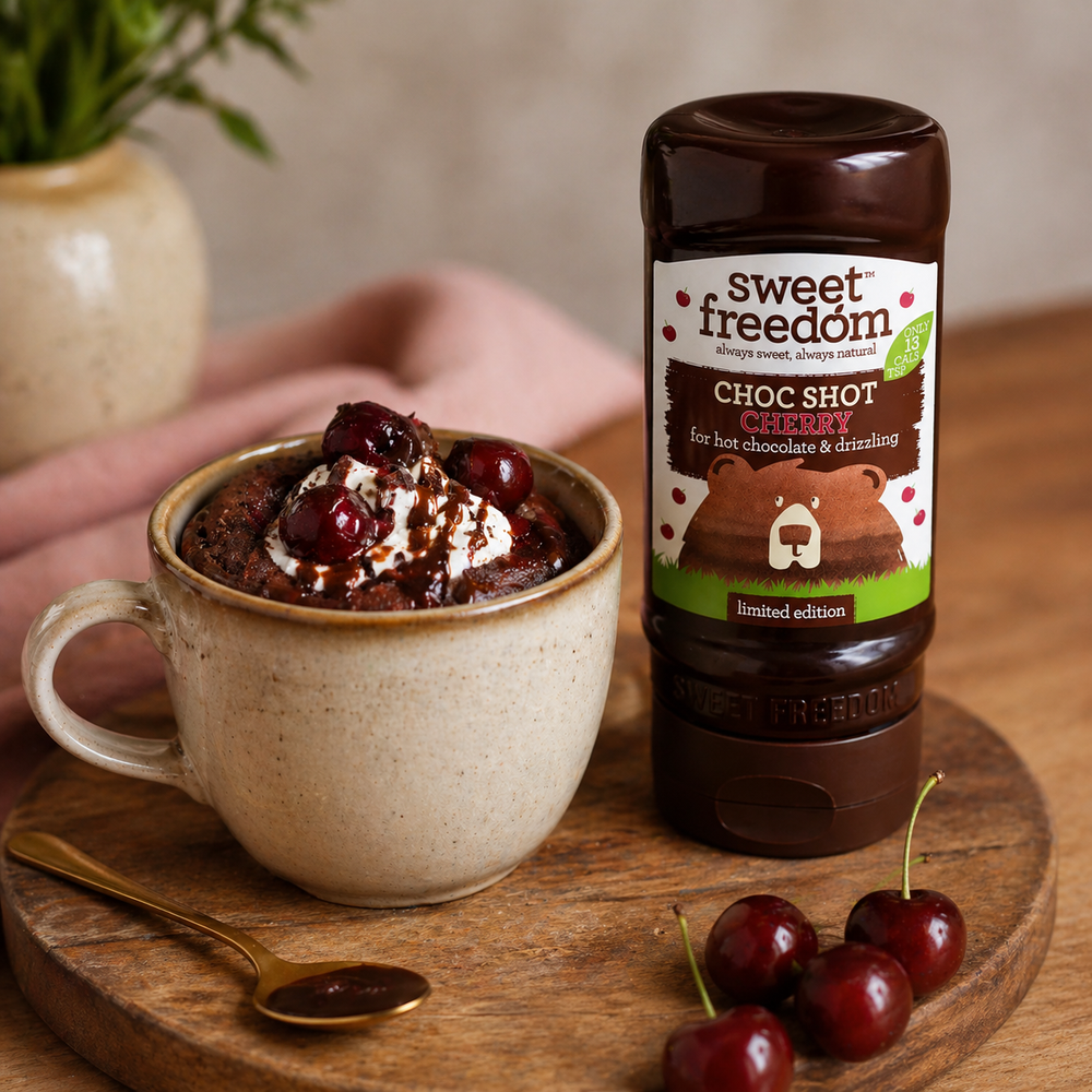 Black Forest Gateau mug cake topped with cherries and a drizzle of Choc Shot Cherry, a bottle of Choc Shot Cherry stands on the board to the right of the mug, and fresh cherries are scattered alongside