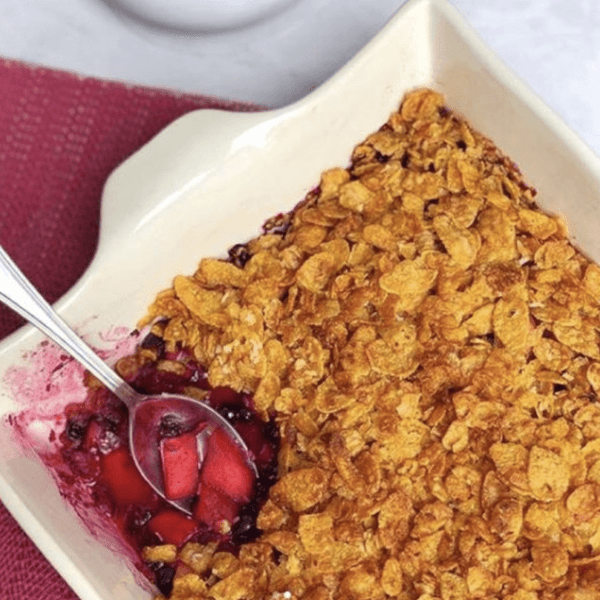 Baking tray filled with delicious apple and berry cornflake crumble, showing a spoon in the top left corner and a portion of the crumble removed.